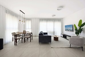A modern living room with a sofa, dining table, chairs, and a TV. - Spacious 4BR Apt l  Sunny Balcony & Mamad (Bat Yam)