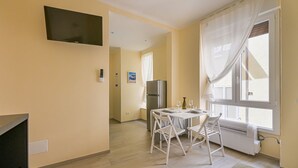 Ein Zimmer mit einem flachbildfernseher an der Wand, einem Esstisch mit Stühlen, einem Kühlschrank und einem fenster mit Vorhängen.