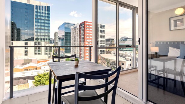 A balcony with a table and chairs, a potted plant, and a view of city buildings. - Newstead Balcony Hideaway | Rooftop Pool | Walk to James St & the Valley (Newstead)