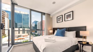 A hotel room with a large bed, a nightstand, a window with a city view, and framed pictures on the wall. - Newstead Balcony Hideaway | Rooftop Pool | Walk to James St & the Valley (Newstead)
