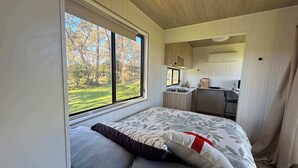 Un dormitorio con un ventanal grande, una cama con ropa de cama floral y una kitchenette al fondo.