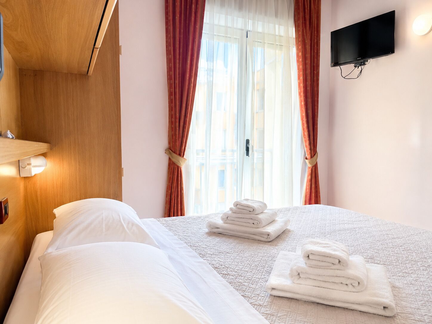 Habitación de hotel con una cama, un televisor de pantalla plana montado en la pared y una ventana con cortinas.