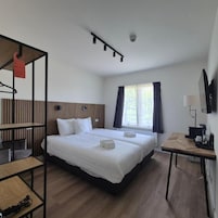 Quarto de hotel com cama, escrivaninha, cadeira, televisão e janela com cortinas.