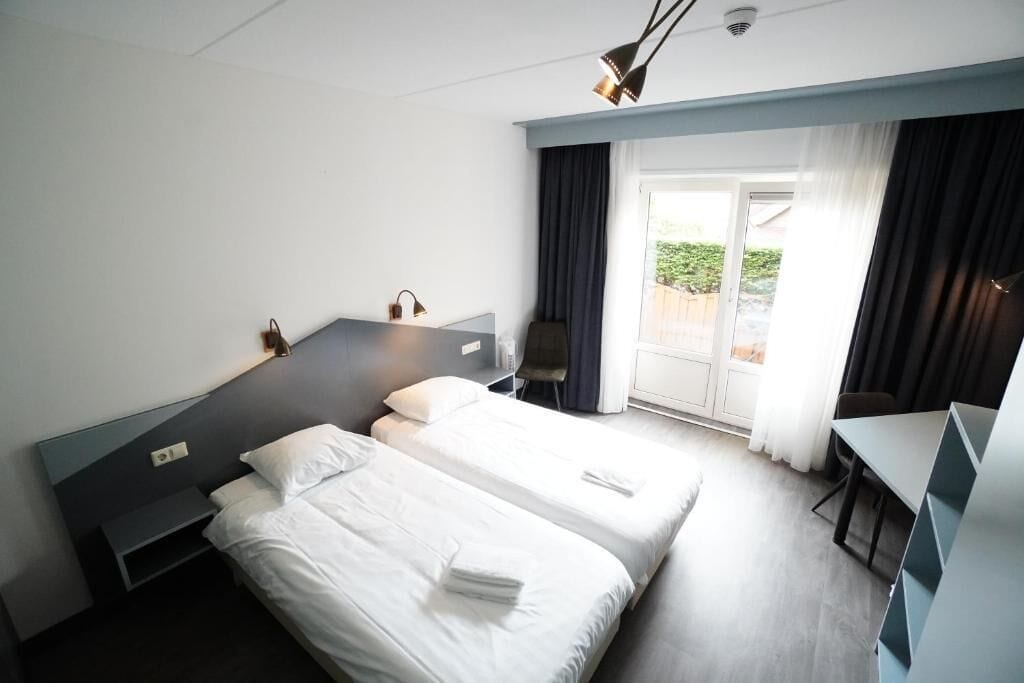 A hotel room with two beds, a desk, a chair, and a view of the outdoors.