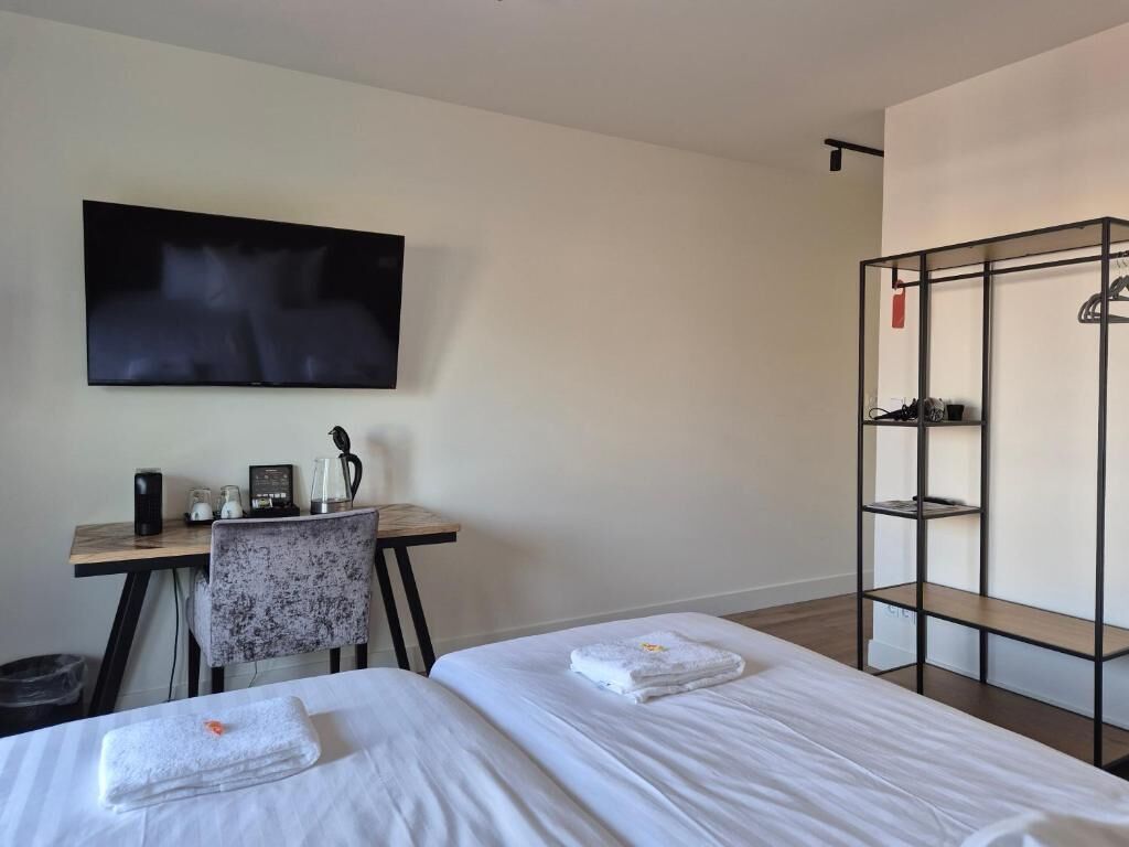 A hotel room with two beds, a desk with a chair, a TV mounted on the wall, and a metal shelf unit.