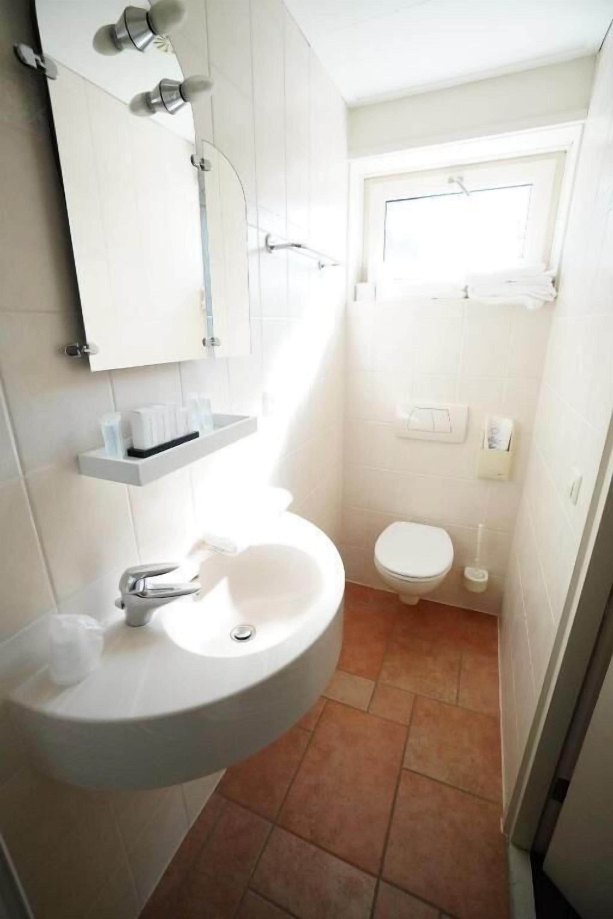 Un baño pequeño con un lavamanos blanco, un espejo, un inodoro y un ventanal sobre el inodoro.