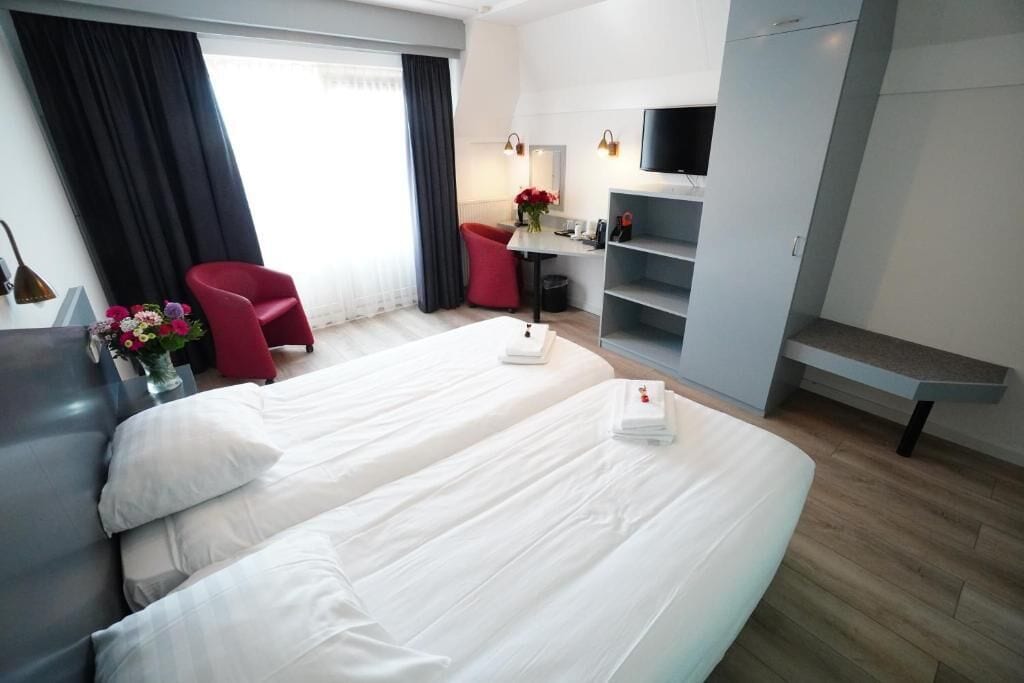 A hotel room with two beds, a desk, a chair, a TV, and a window with curtains.