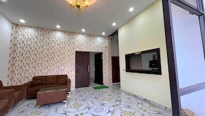 A room with patterned wallpaper, a sofa, a coffee table, a television mounted on the wall, and a door leading to another room. - Sunrise Prime (Ranchi)