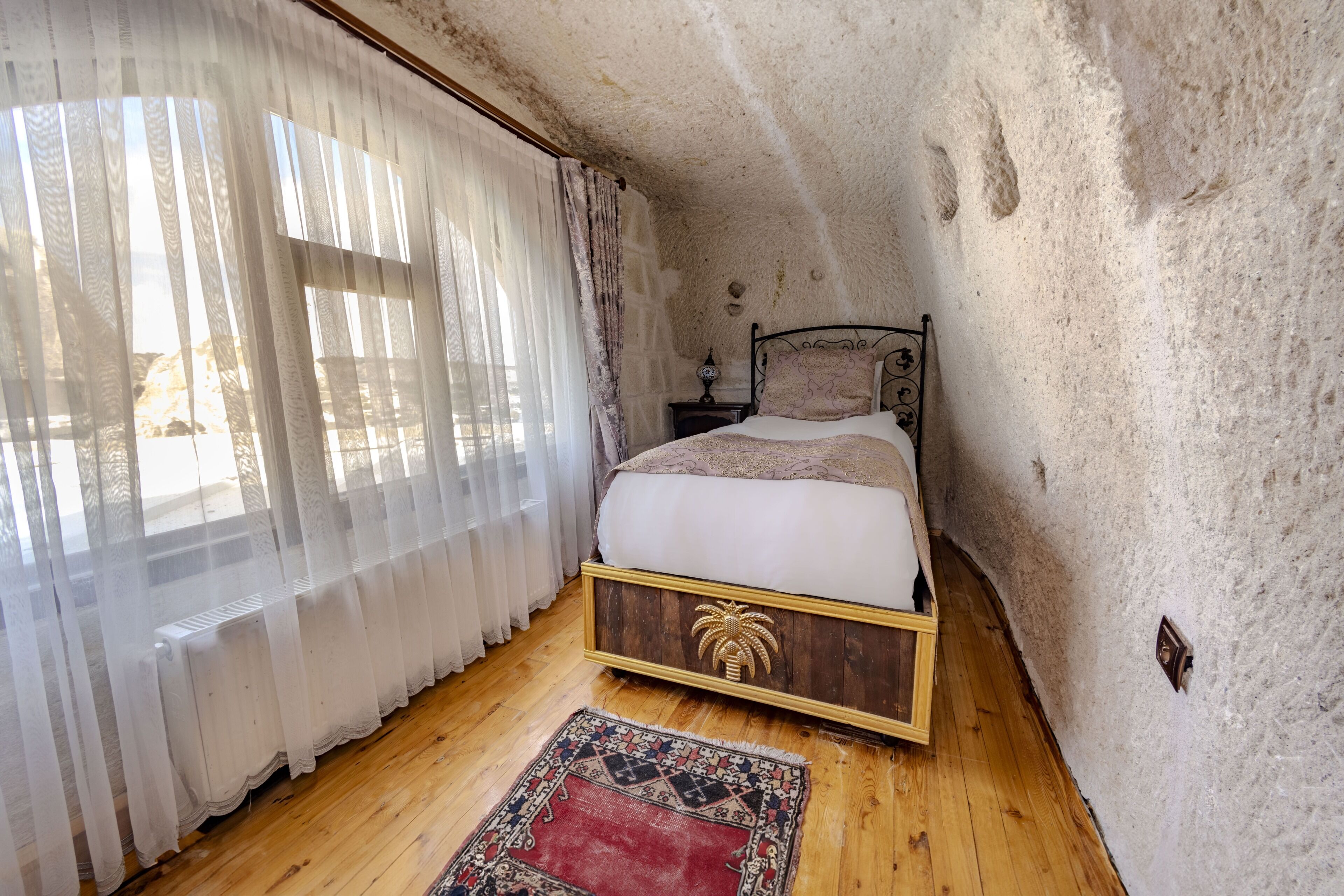 A bedroom with a wooden floor, a bed with a headboard, a nightstand, a lamp, and a window with sheer curtains.