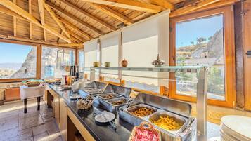 A buffet setup with various food items and utensils, a wooden ceiling, and a view of the outdoors.
