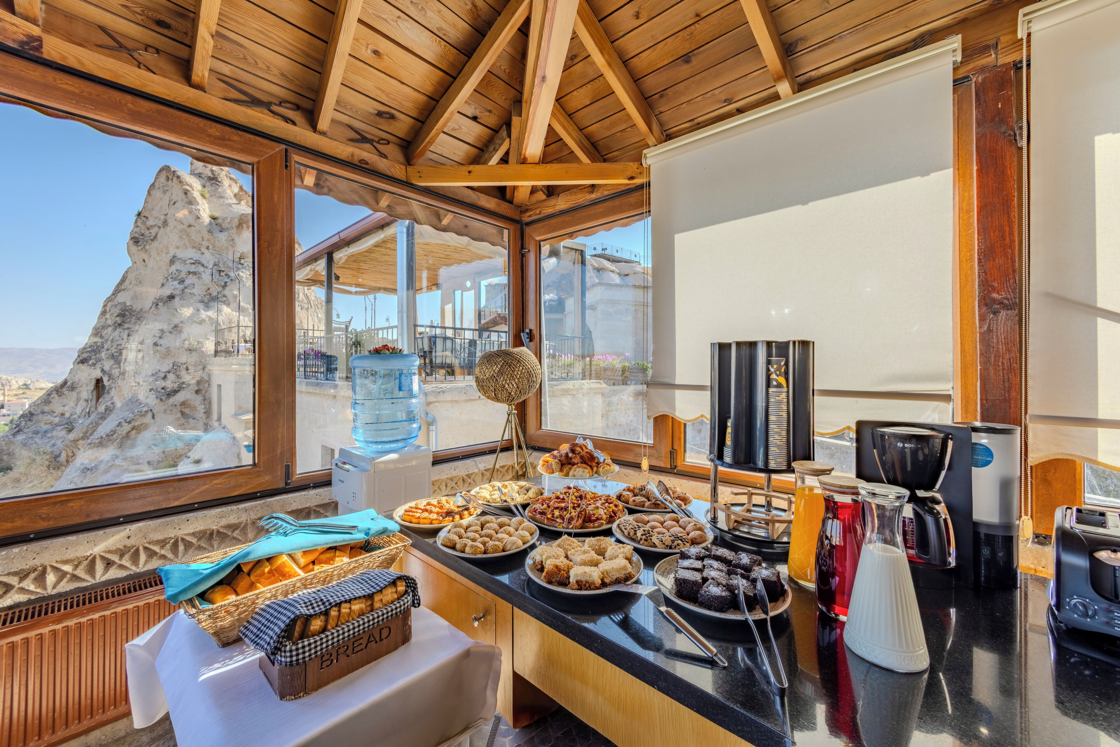 Un bufé de desayuno con una variedad de pasteles y pan, un dispensador de agua y una máquina de café en una habitación con vigas de madera y vistas a un paisaje rocoso.