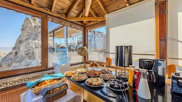 Un bufé de desayuno con una variedad de pasteles y pan, un dispensador de agua y una máquina de café en una habitación con vigas de madera y vistas a un paisaje rocoso.
