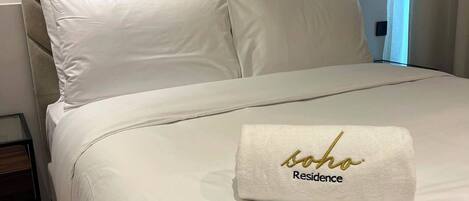 Una cama bien tendida con un cabecero color beige, ropa de cama blanca y una toalla con la inscripción 'soho Residence'.