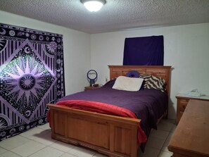 A bedroom with a bed, a wooden headboard, a bedside table, a nightstand, a wall tapestry, and a ceiling light. - A Clean, Well-lighted Place:
Tropical Holiday Haven~Where Groove Gets Got Again (Holiday)