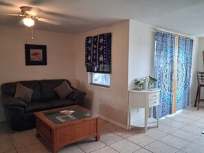A living room with a dark sofa, a wooden coffee table, a ceiling fan, a window with curtains, and a small white cabinet. - A Clean, Well-lighted Place:
Tropical Holiday Haven~Where Groove Gets Got Again (Holiday)