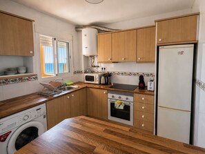 A compact kitchen with wooden cabinets, a built-in washing machine, a microwave, a stove, and a refrigerator. - Seagull Lookout (Mojácar)
