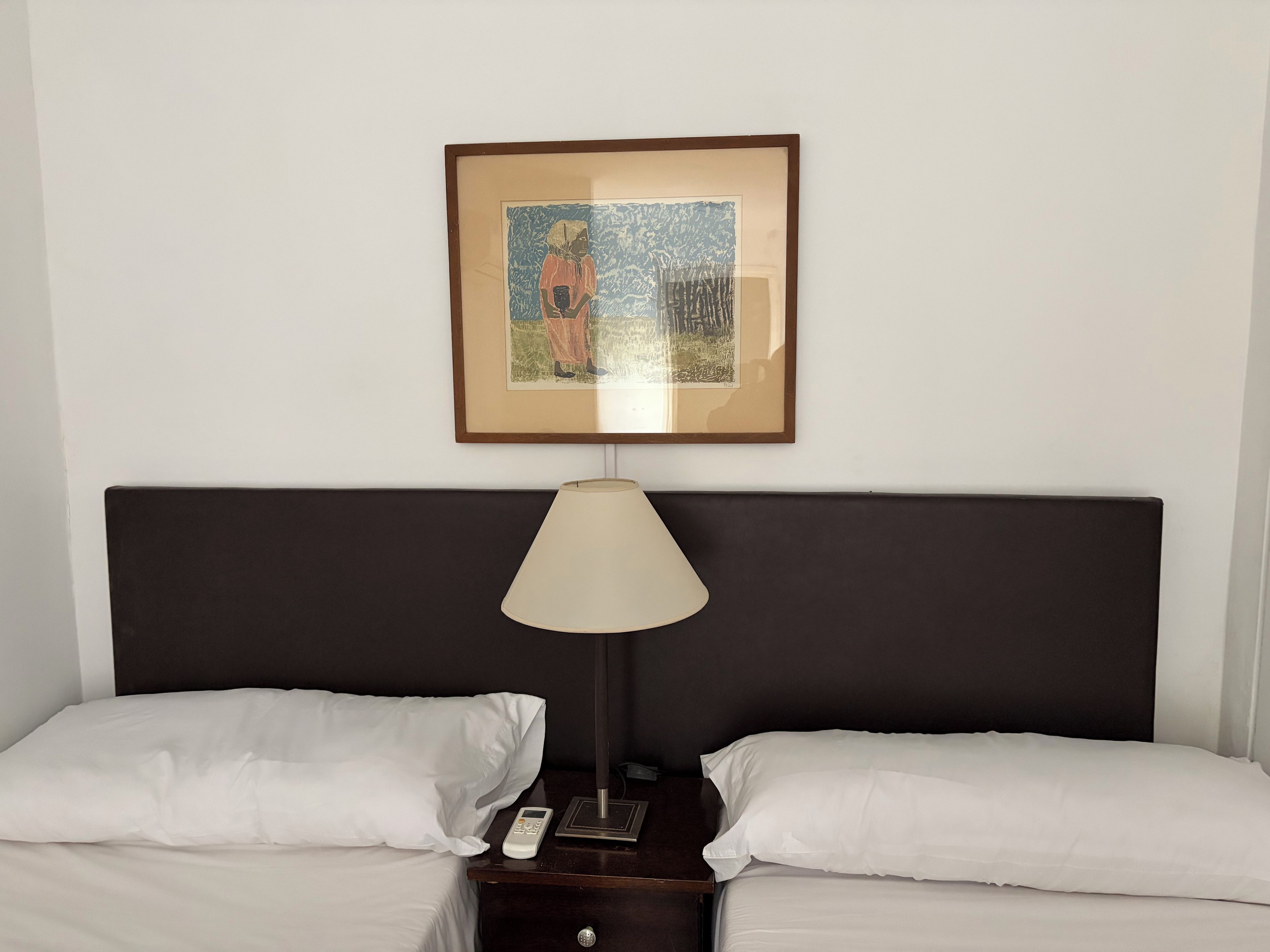 Et hotelværelse med en seng, to puder, et natbord med en lampe, en fjernbetjening og et billede i ramme på væggen.