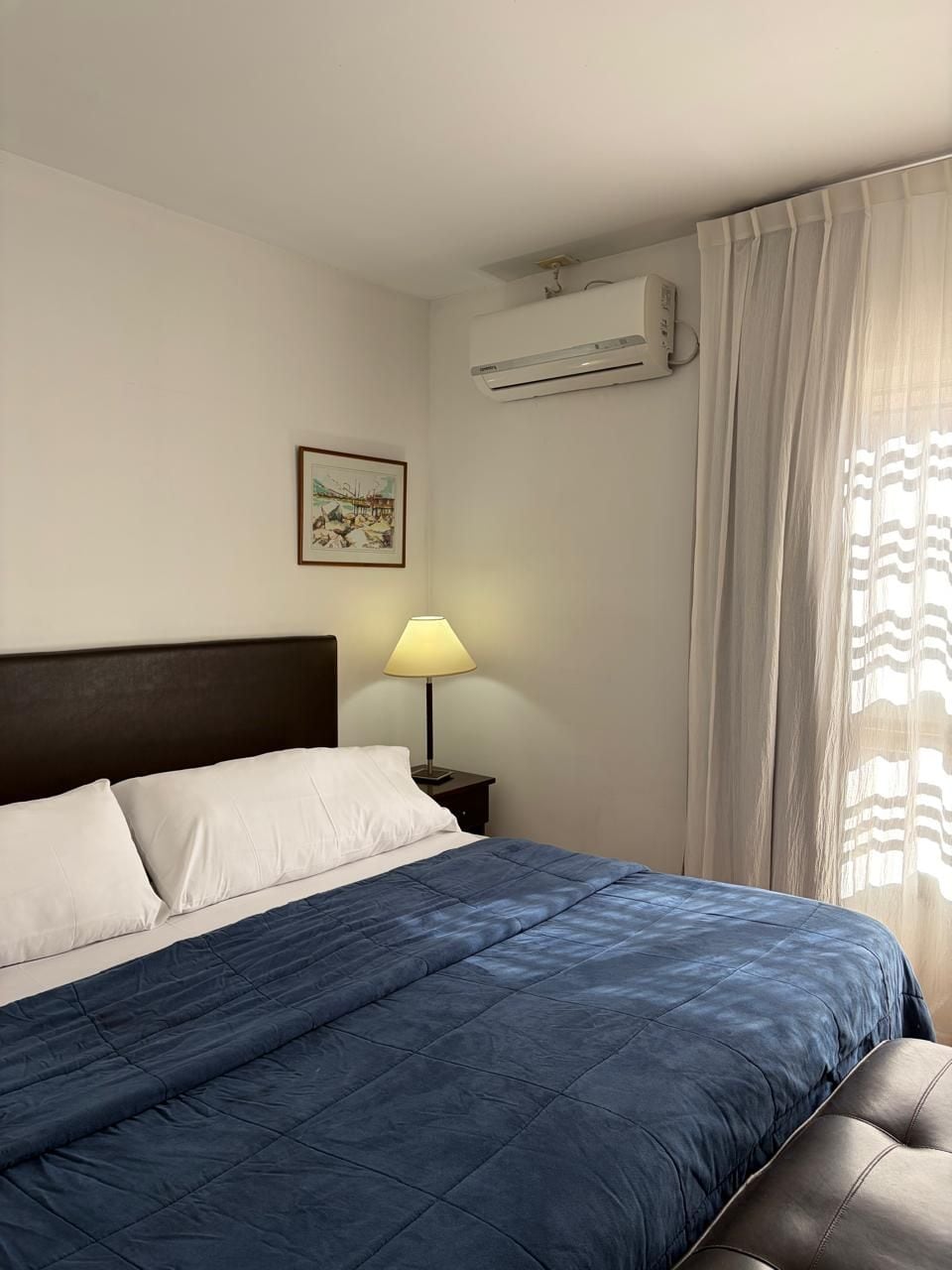 A hotel room with a bed, a nightstand, a lamp, an air conditioning unit, and a window with curtains.
