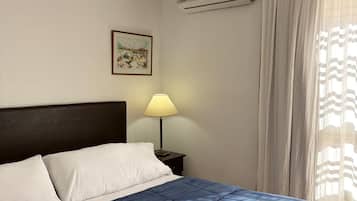 A hotel room with a bed, a nightstand, a lamp, an air conditioning unit, and a window with curtains.