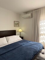 Et hotelværelse med seng, natbord, lampe, klimaanlæg og et vindue med gardiner.