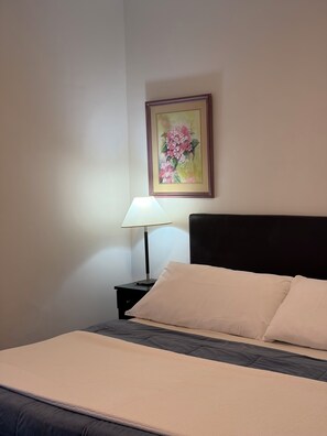 Una cama bien hecha con un almohadón blanco y un cabecero oscuro, una lámpara de noche y un cuadro con flores en la pared.