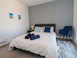 Una cama bien hecha con toallas enrolladas, una silla azul y una mesita de noche con lámpara y marco de foto.