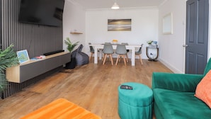 Una sala de estar moderna con un televisor de pantalla plana, un comedor con una mesa y sillas blancas, un sofá verde y piso de madera.