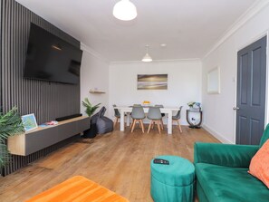 A modern living room with a flat-screen TV, a dining area with a white table and chairs, a green sofa, and a wooden floor. - Ty Haul (Rhosneigr)