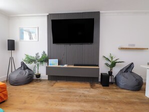Ein modernes Wohnzimmer mit einem an der Wand mit vertikalen Streifen befestigten Flachbildfernseher, einem Holzboden, zwei grauen Sitzsäcken, einem kleinen Tisch mit einer Karte und Büchern sowie einer Stehlampe.