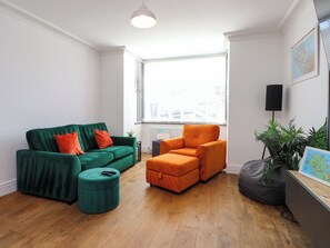 Ein Wohnzimmer mit einem grünen Sofa, einem orangefarbenen Sessel, einem türkisblauen Hocker, einer schwarzen Stehlampe und einer Karte auf einem Schreibtisch.