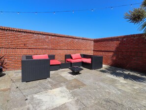 Une terrasse dotée de meubles en osier et de coussins rouges, d’un mur en briques et de guirlandes lumineuses.