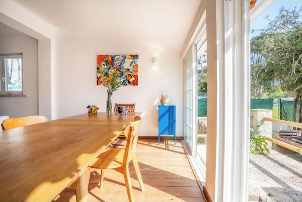 A dining area with a wooden table and chairs, a colorful abstract painting on the wall, and a view of the outdoors through large windows.