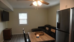 A compact kitchen with a dining table, chairs, a refrigerator, a ceiling fan, a television, and a window.