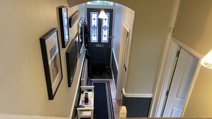 Un pasillo estrecho con una escalera, una pequeña mesa con lámpara y una puerta que da a una habitación con ventana.