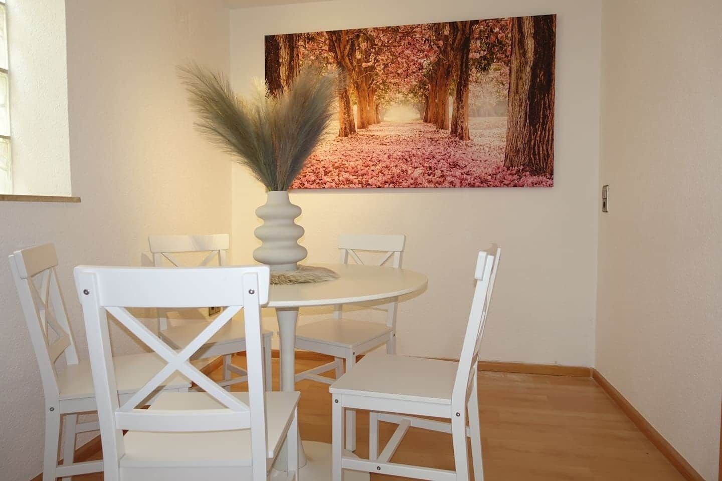A dining area with a round table, white chairs, and a vase with decorative foliage.