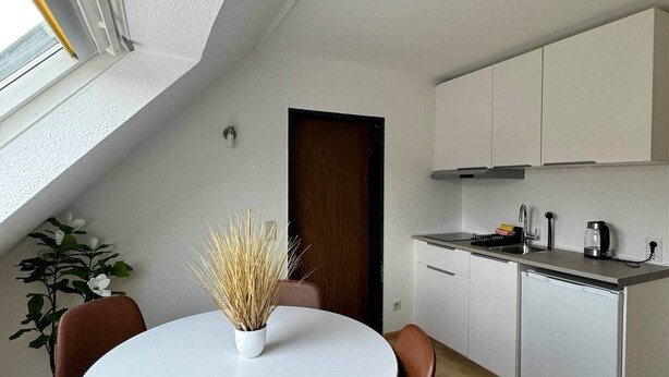 A modern kitchen with white cabinets, a built-in oven, and a dining area with a white table and brown chairs. - FeWo Moments - 2 min to E-Park (Rust)