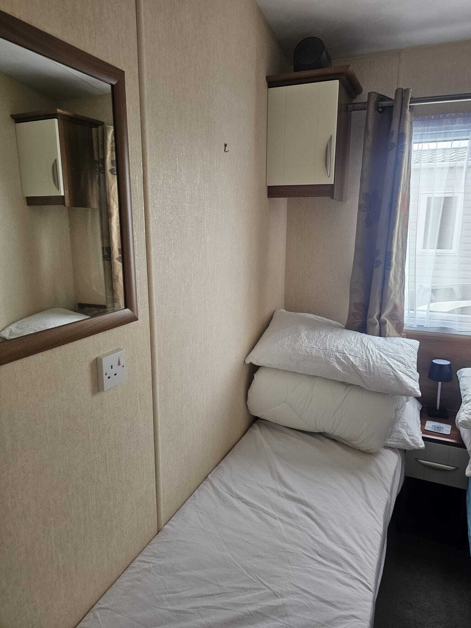 Ett litet hotellrum med en enkel säng, ett nattduksbord med en lampa, en spegel och ett fönster med gardiner.