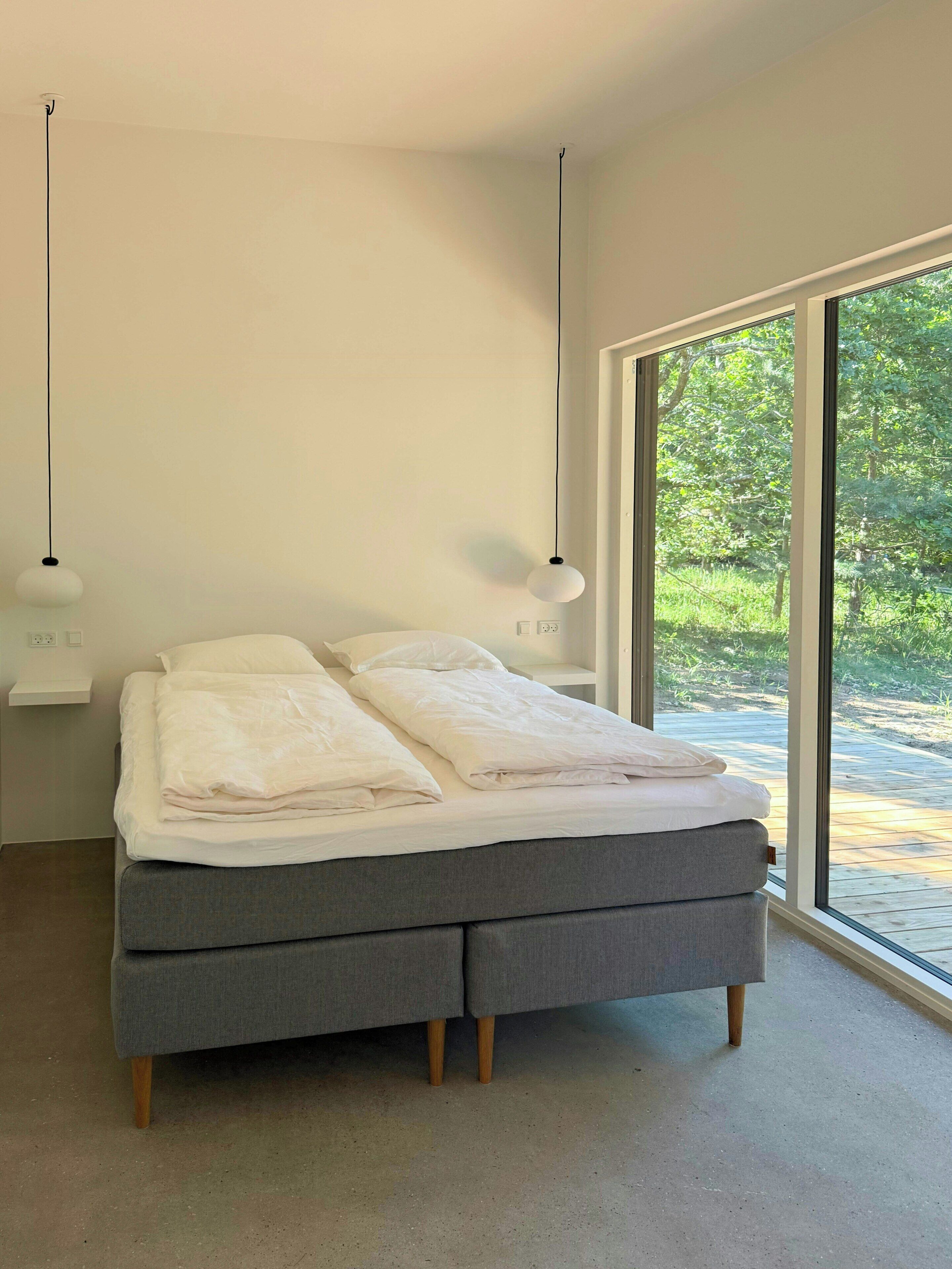 A modern bedroom with a large bed, two pendant lights, and a view of a forest through a large window.