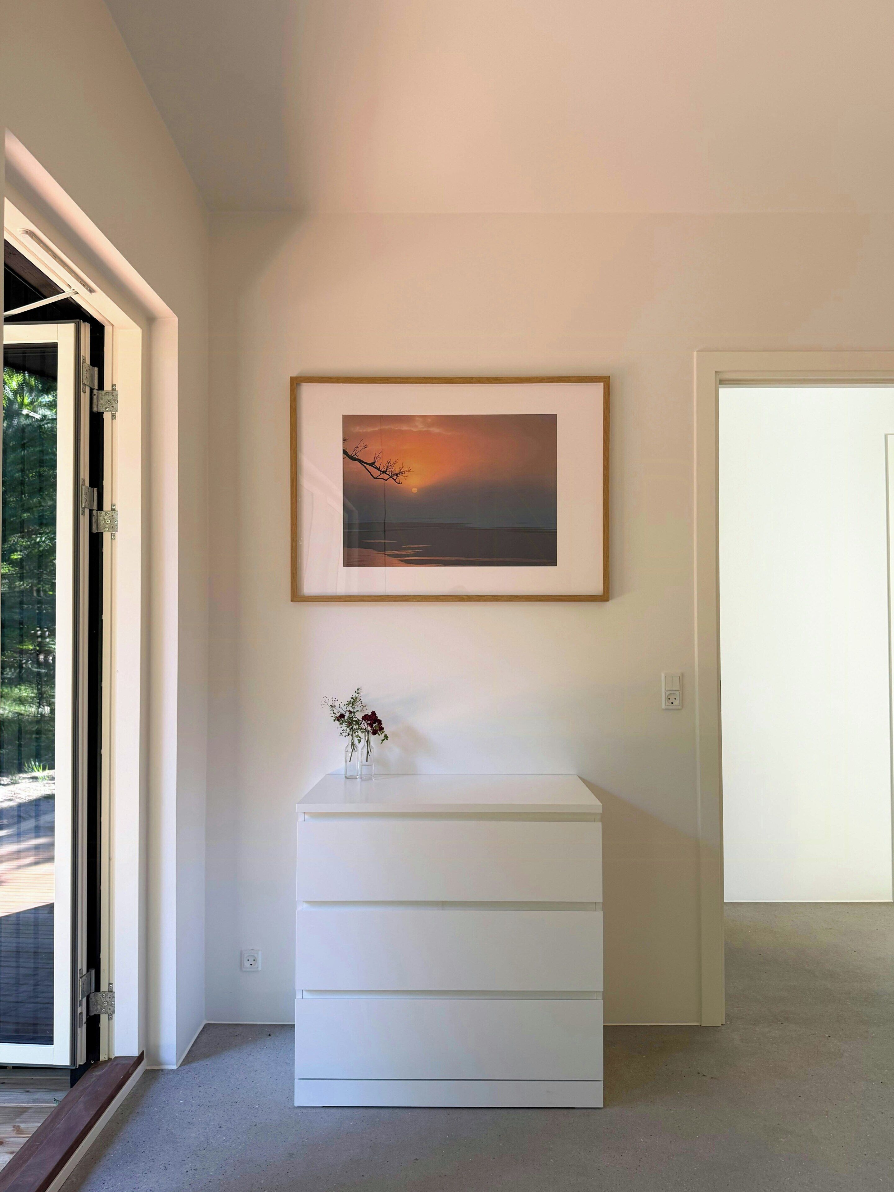 Una habitación con un tocador blanco, un cuadro enmarco de un atardecer y un pequeño florero sobre el tocador.
