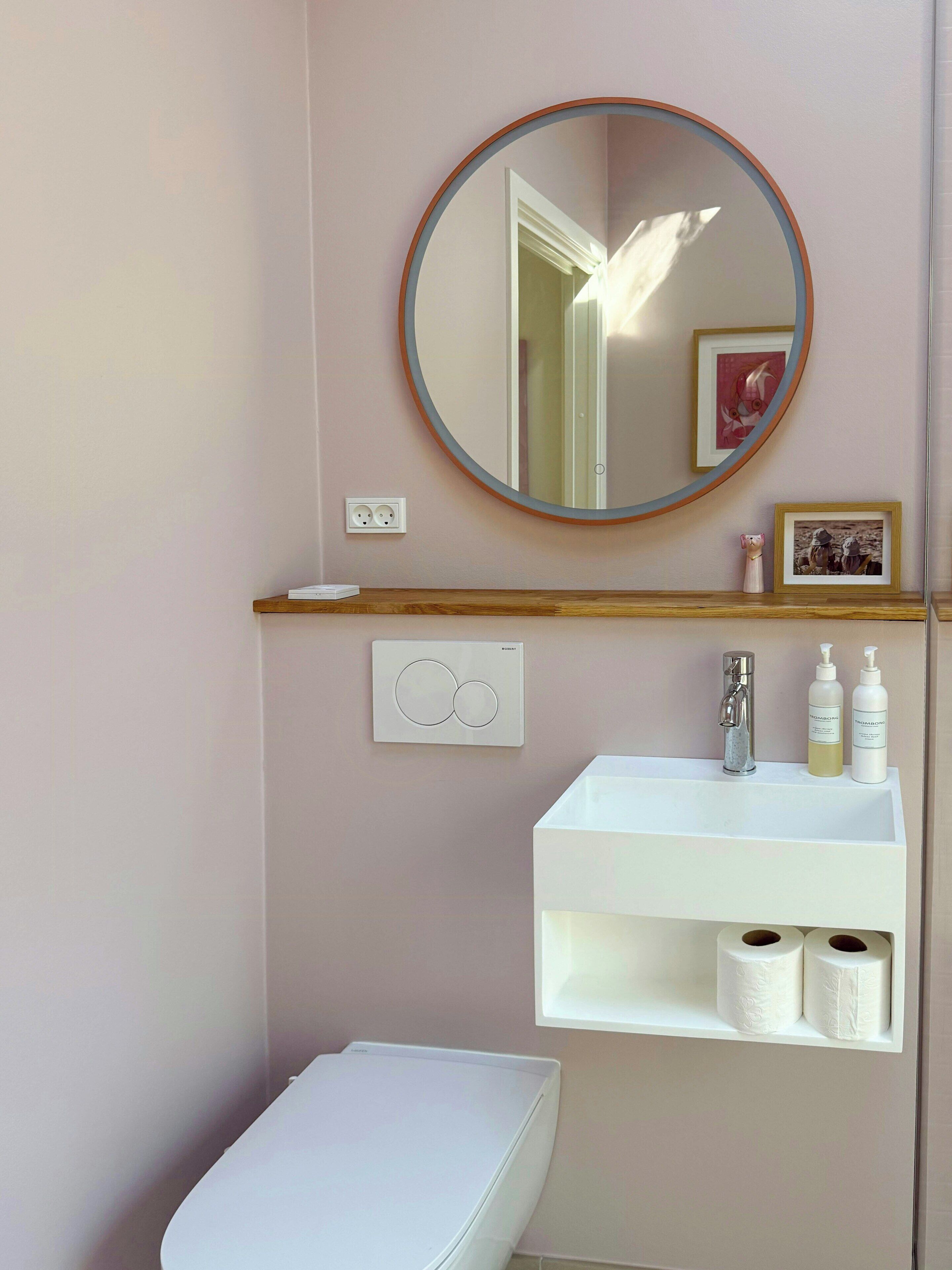 A bathroom with a round mirror, a white sink, a toilet, and a shelf with toiletries.