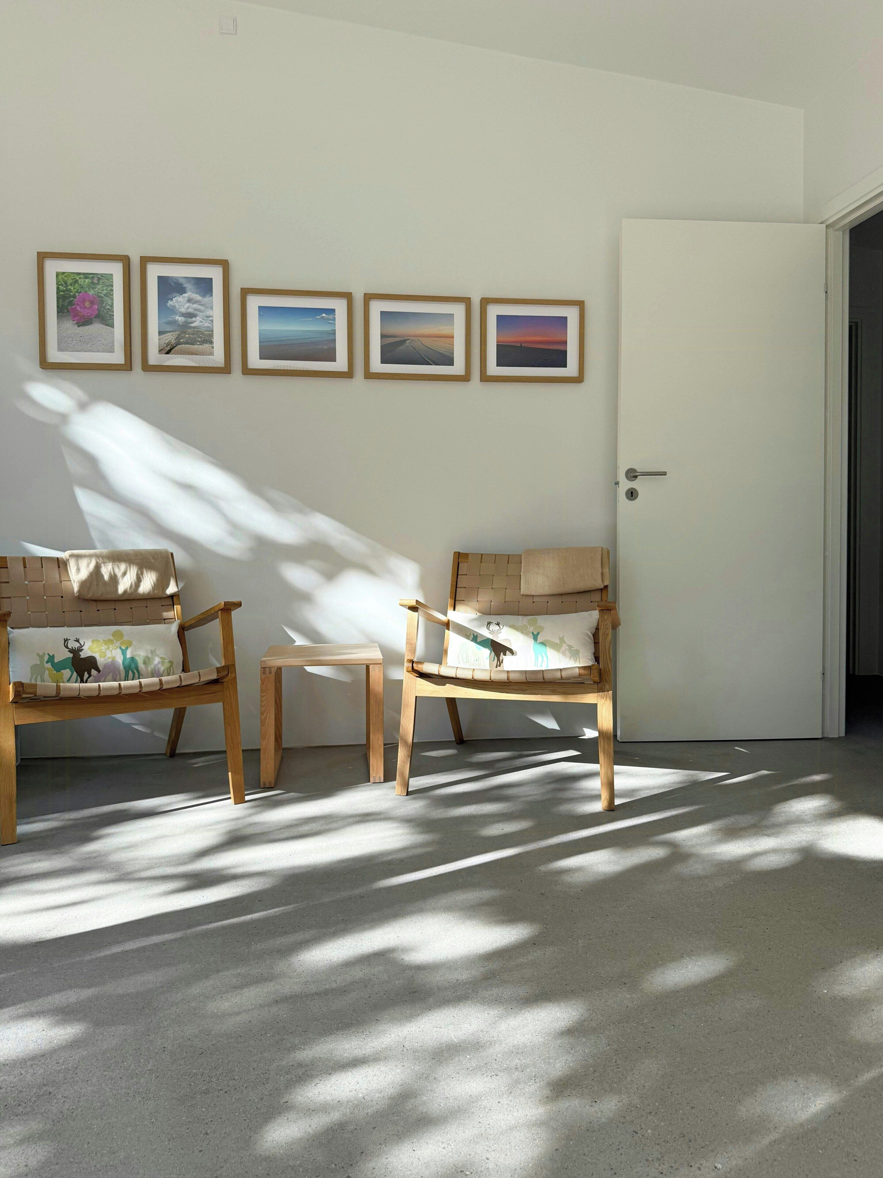 A room with two wooden chairs, a small table, and a door. There are four framed pictures on the wall.