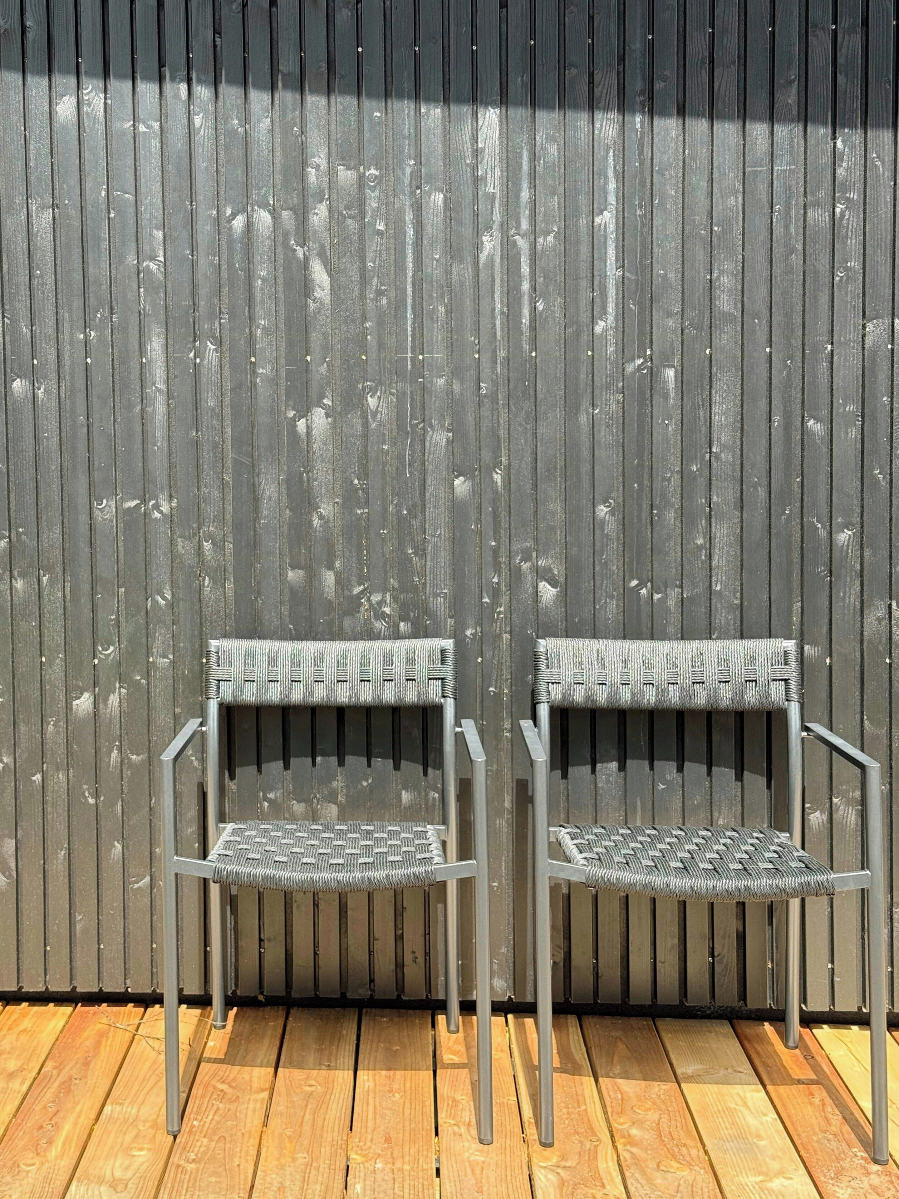 Dos sillas de metal con asientos y respaldos tejidos, ubicadas frente a una pared de metal corrugado.