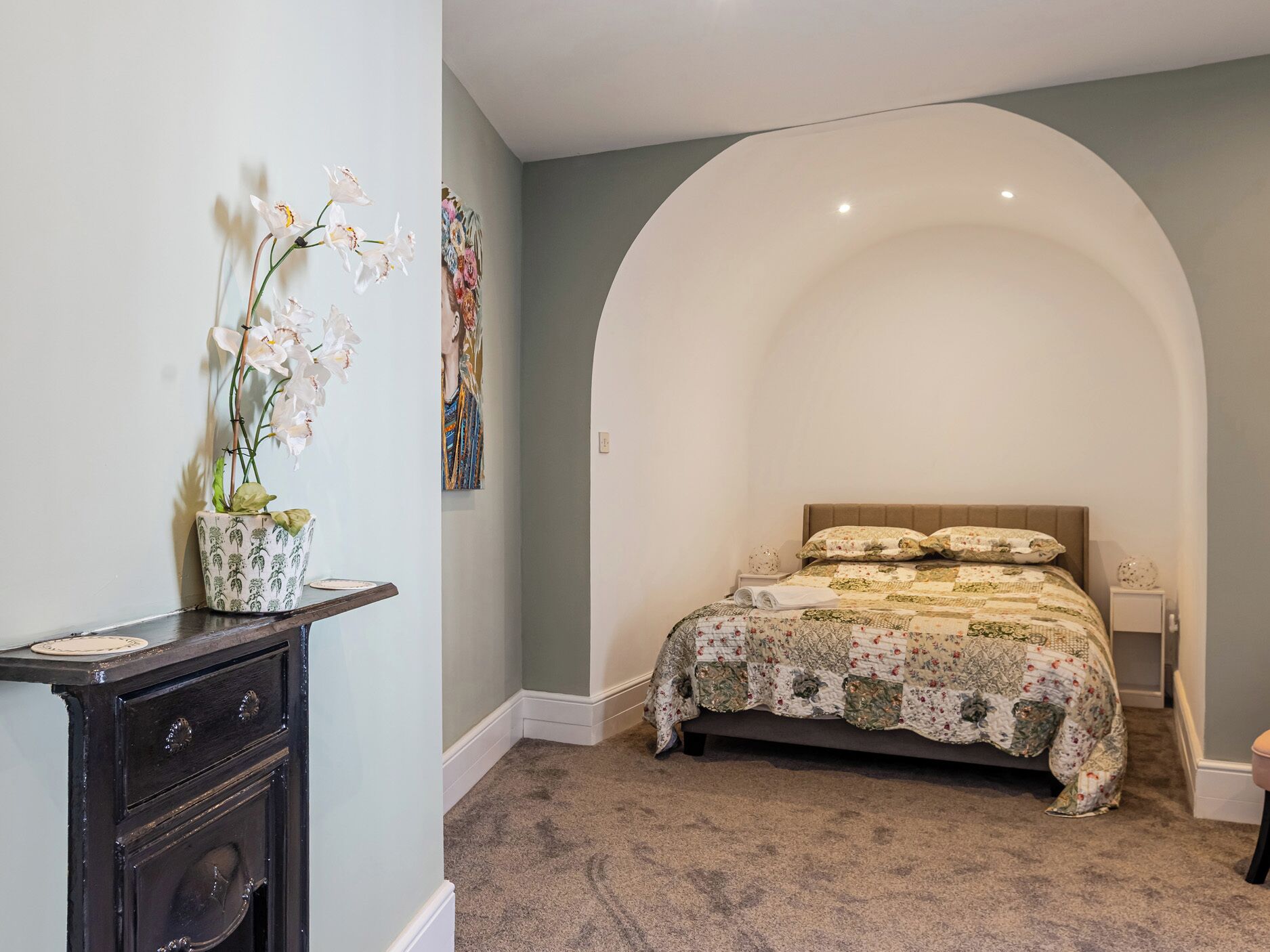 A bedroom with a large arched window, a bed with a floral quilt, a nightstand, a dresser, and an orchid plant.