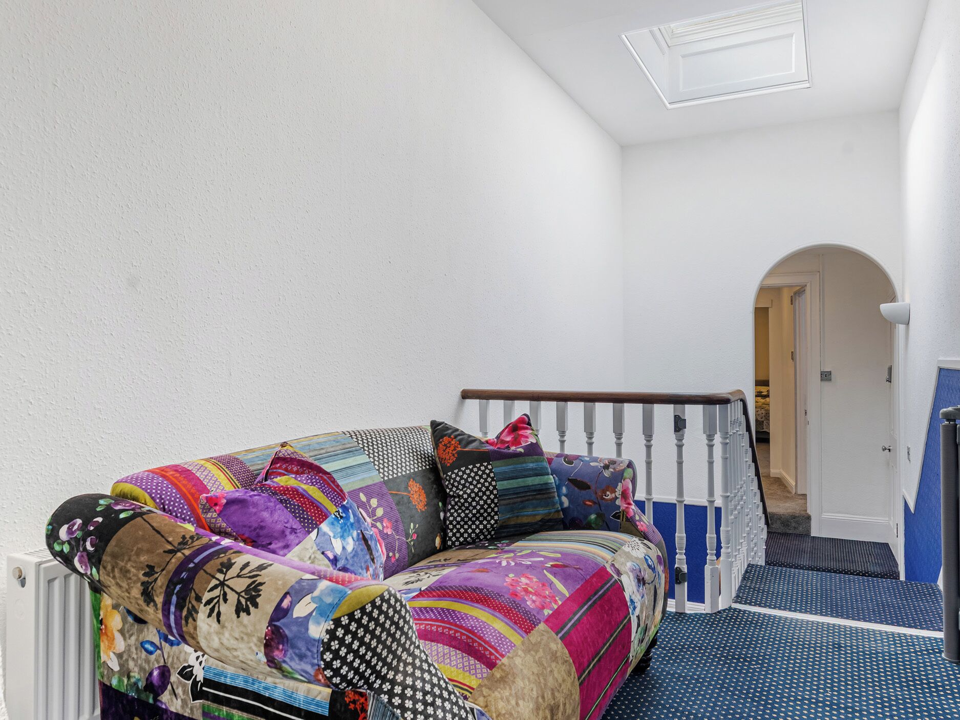 Une pièce aux murs blancs, avec un canapé coloré, un escalier en bois et un sol à motifs bleus.