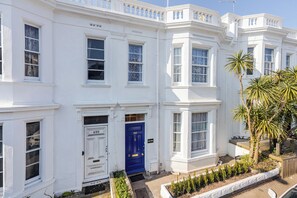 A white townhouse with a blue door, numbered 493, and a small garden with plants. - Palm Villa (Torquay)