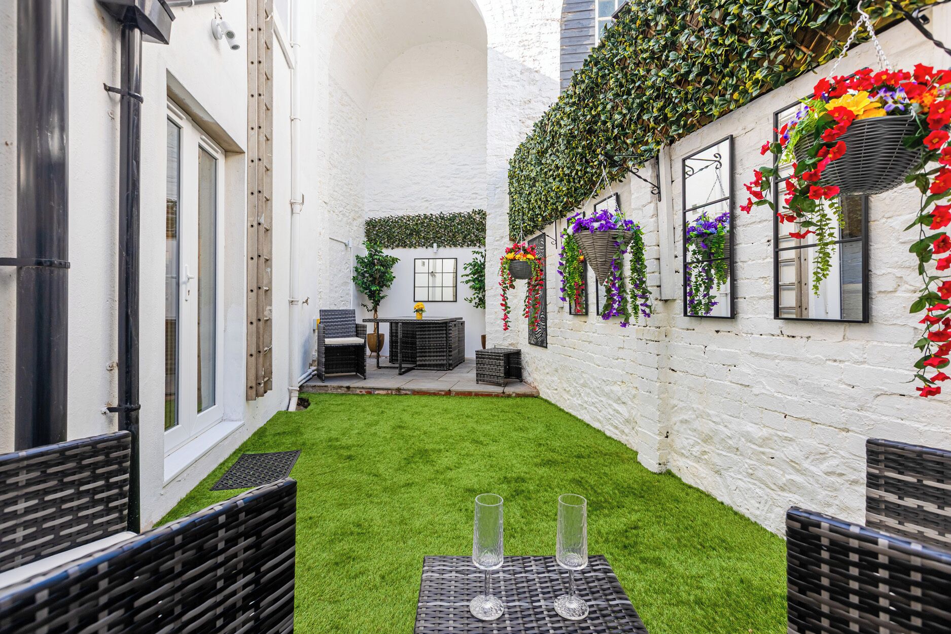 Une petite cour étroite, avec un mur en briques blanches, un petit espace aménagé avec une table et des chaises, et des jardinières suspendues.
