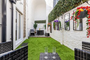 Ein schmaler Innenhof mit einer weißen Backsteinmauer, einer kleinen Sitzecke mit Tisch und Stühlen sowie hängenden Blumenkörben.
