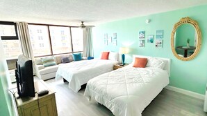 Ein Hotelzimmer mit zwei Betten, einem Schreibtisch mit Computer, einem Sessel, einem runden Tisch mit Lampe und einem großen Spiegel.