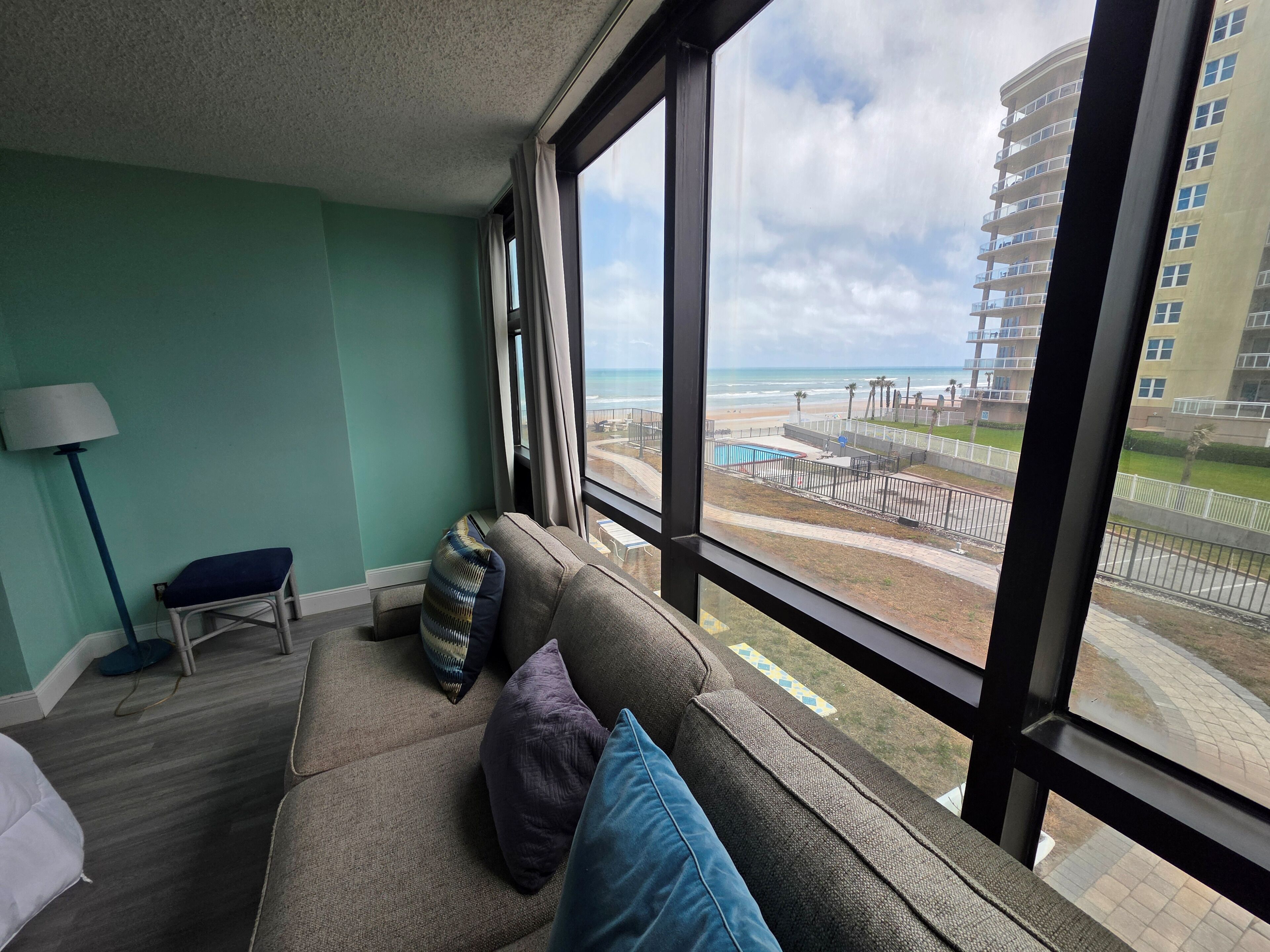 Ein Zimmer mit großem Fenster, einer Couch mit Kissen, einer Lampe und Blick auf Strand und Gebäude.