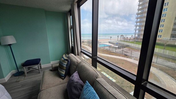 Ein Zimmer mit großem Fenster, einer Couch mit Kissen, einer Lampe und Blick auf Strand und Gebäude.